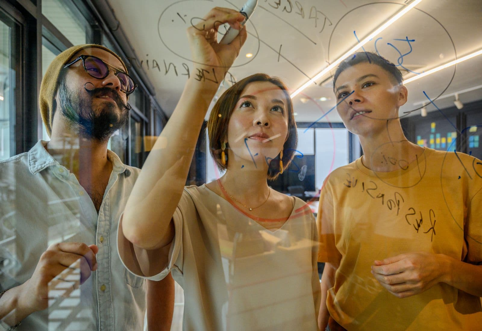 Três pessoas desenhando um diagrama num quadro de vidro.